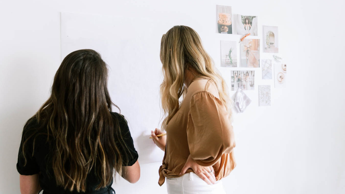 Two women facing a wall writing on a white board.